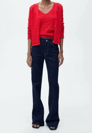 Roter, gestrickter Cardigan über einem passenden ärmellosen Top, kombiniert mit ausgestellten dunklen Denim-Jeans, die eine glatte Textur und klassische Passform zeigen.