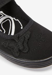 Zapato de tela negra con una textura suave, que presenta un bordado de mariposa y sutiles detalles de costura. Punta redondeada y tira elástica.