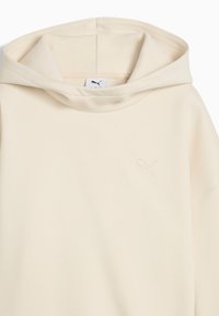 Sudadera color crema con una textura suave y lisa y un cuello alto. Presenta un pequeño logo de Puma en relieve en el pecho.