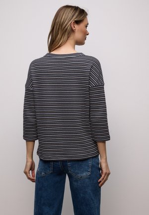 Femme aux cheveux châtain clair portant un haut à manches longues à motifs bleu marine et beige ainsi qu'un jean bleu, vue de dos sur un fond uni.