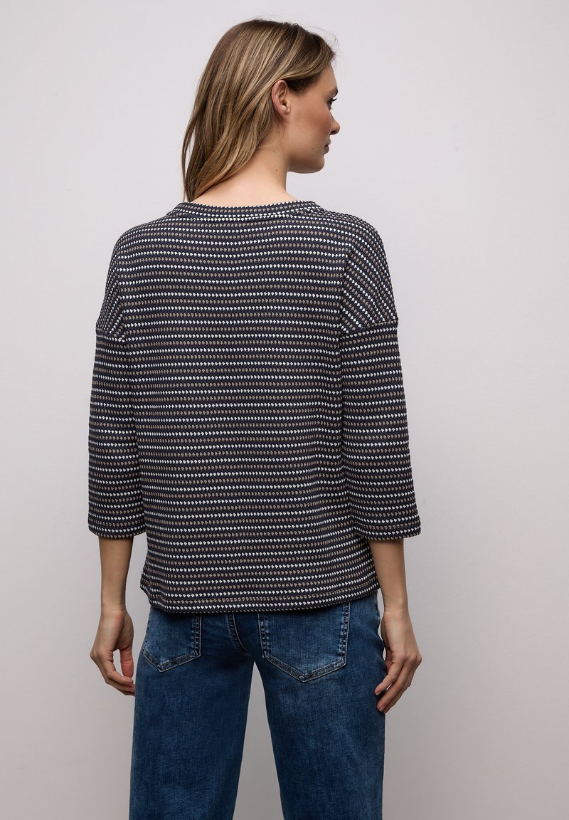 Femme aux cheveux châtain clair portant un haut à manches longues à motifs bleu marine et beige ainsi qu'un jean bleu, vue de dos sur un fond uni.