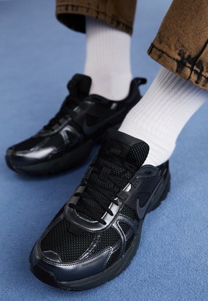 Osoba nosząca czarne sneakersy Nike z białymi skarpetkami i brązowymi spodniami stojąca na niebieskiej powierzchni.