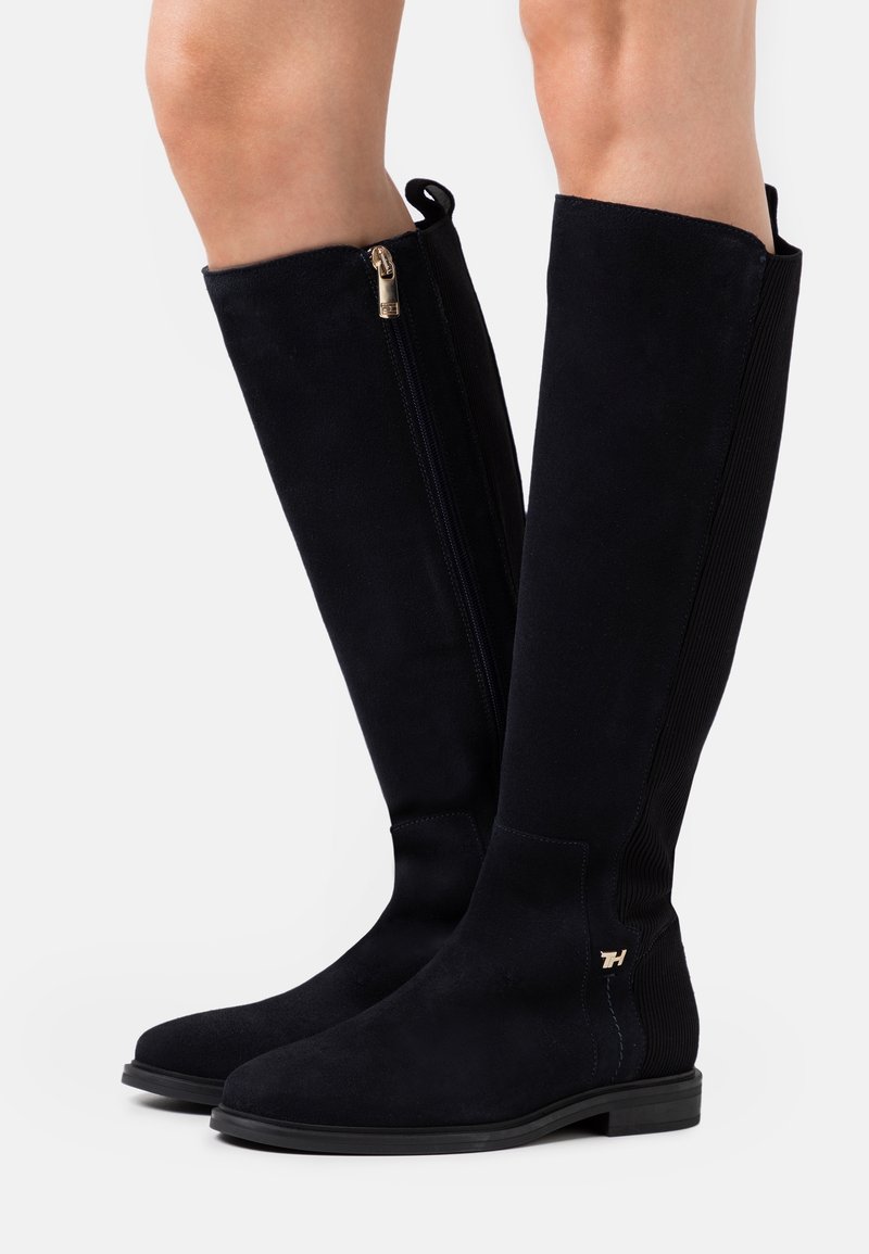 Bottes noires en daim montant jusqu'au genou avec fermeture éclair latérale, talon bas, panneau élastique à l'arrière et petit logo doré près de la cheville sur un fond neutre.