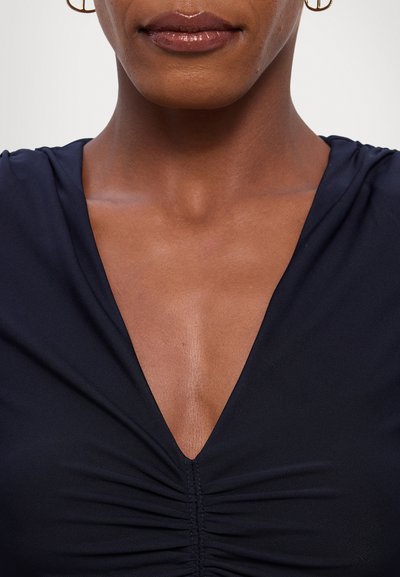 Navy blue v-neck top with ruched detailing at the center, featuring a smooth texture and fitted shape. Gold hoop earrings visible.