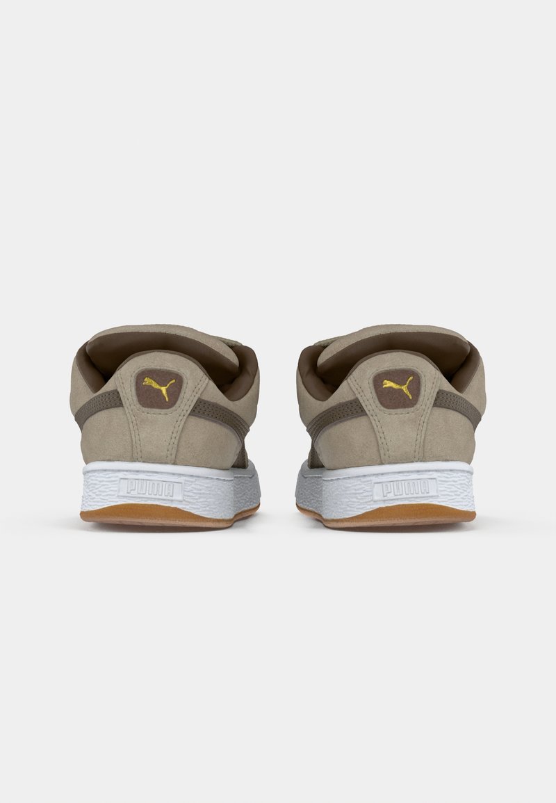 Zapatillas Puma con una parte superior de gamuza beige y acentos oscuros. Suela texturizada blanca y suela de goma marrón. Presenta un logo en la parte trasera.
