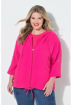 Uśmiechnięta kobieta o blond włosach, nosząca duże złote kolczyki-koła, jasnoróżową kurtkę na zamek i niebieskie jeansy z kwiatowym haftem.