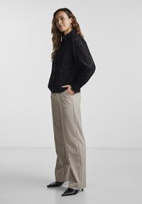 Black embroidered blouse with long puff sleeves, paired with beige high-waisted trousers and pointed black heels. Smooth fabric, intricate pattern.