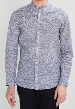 Homme portant une chemise à manches longues boutonnée avec un petit motif floral noir et blanc et un pantalon noir sur fond uni.