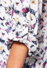 Chemise à motif floral en blanc, avec des couleurs incluant le rose, le violet et le jaune. Elle dispose d'un poignet retroussé avec un détail bouton décoratif.