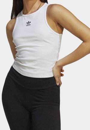 Kobieta ubrana w białą, bezrękawnikową koszulkę Adidas i czarne legginsy, stojąca z jedną ręką na biodrze na tle jednolitego tła.