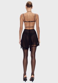 Kobieta stoi tyłem, ubrana w czarny stanik na cienkich ramiączkach, przezroczystą czarną spódnicę, czarne pończochy z szwem oraz czarne sandały na wysokim obcasie.