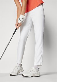 Λευκά παντελόνια γκολφ με φερμουάρ στο πλάι, συνδυασμένα με ανοιχτόχρωπα παπούτσια γκολφ και ένα λευκό γάντι. Το επάνω μέρος έχει πορτοκαλί απόχρωση και υφή σχεδίου.