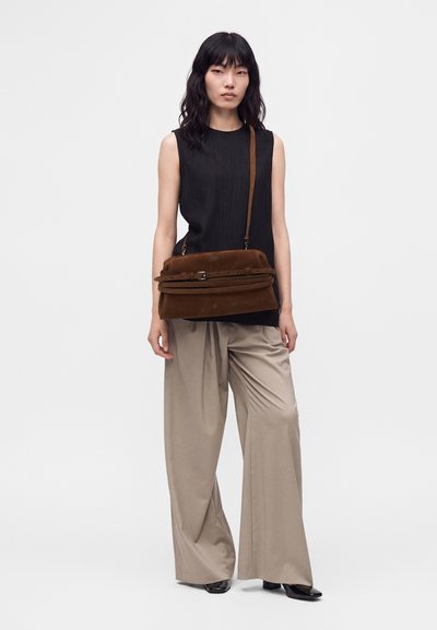 Femme avec un haut noir sans manches et un pantalon large beige, portant un grand sac marron en bandoulière, debout devant un fond uni.