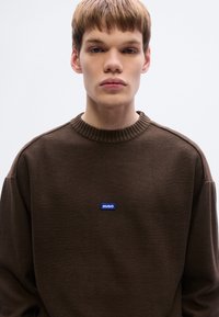 Sweat-shirt marron avec un col côtelé et des coutures tombantes aux épaules. Comprend un patch logo rectangulaire bleu avec le texte blanc "HUGO" sur la poitrine.