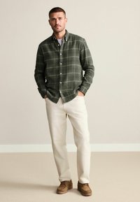 Camisa de cuadros verde con cuello, combinada con pantalones beige claros y zapatos marrones. La camisa tiene una textura suave y un corte regular.