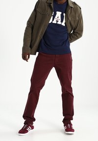 Homme portant une veste vert olive, un sweat-shirt bleu marine avec un logo blanc, un pantalon bordeaux et des baskets assorties, debout sur un fond blanc.