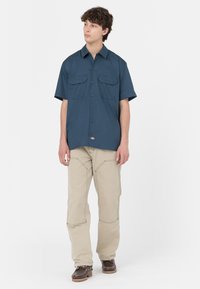 Camicia a maniche corte con colletto in tessuto blu scuro, con due tasche sul petto. Abbinata a pantaloni beige e scarpe marroni. Design semplice, minimalismo nei dettagli.