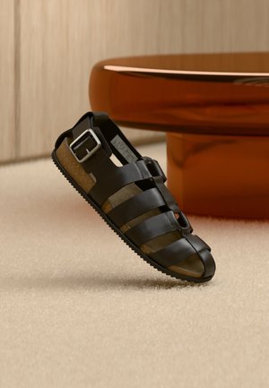 Sandalia de cuero negro con múltiples tiras y una hebilla plateada, mostrada sobre una alfombra beige frente a una mesa redonda de madera.