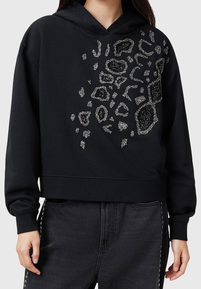 Sweat à capuche court noir avec un imprimé léopard en strass argenté sur la poitrine, porté avec un pantalon noir orné de clous argentés sur les côtés.