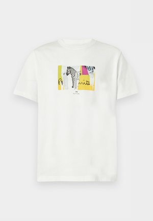 Mand iført en hvid grafisk t-shirt med et zebra-design, blå vide jeans og beige loafers står mod en hvid baggrund.