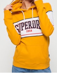 Sudadera amarilla con capucha y bolsillo frontal, con una franja horizontal blanca y roja que lleva el texto "SUPERDRY 1954" en el centro.