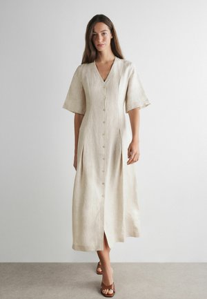 Kvinde står iført en beige midi-kjole med knapper, albuelange ærmer og brune åbne sandaler mod en ensfarvet hvid baggrund.