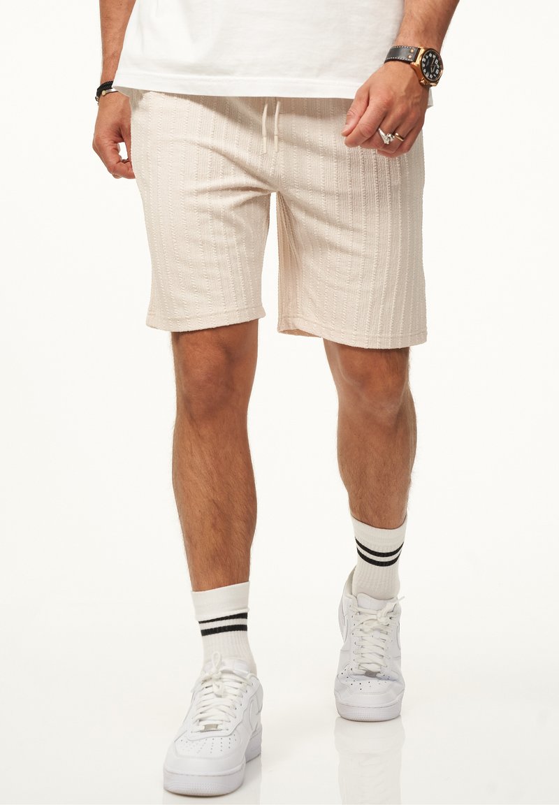 Helle beige Shorts mit strukturierten vertikalen Streifen, elastischem Bund mit Kordelzug und Seitentaschen, kombiniert mit weißen Sneakers und gestreiften Socken.