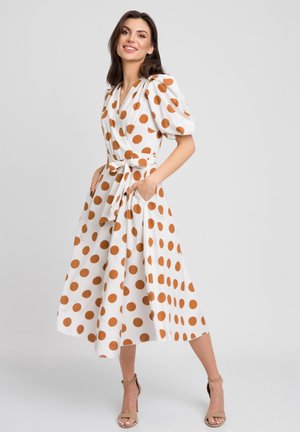 Sukienka w kropki w kolorze białym i rdzawym, z bufiastymi rękawami, fasonem kopertowym, ściśniętą talią i kieszeniami po bokach. Gładki materiał i długość średnia.
