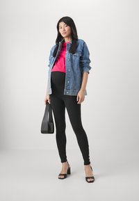 Denimjakke, rosa topp, svarte leggings med gullknappr detaljer, og svarte høyhælte sandaler. Modellen holder en svart veske.