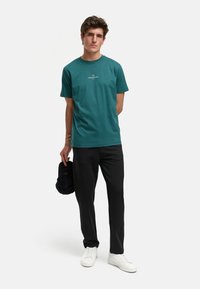 Tyrkise bomull t-skjorte med rund hals og korte ermer, med en hvit logo. Kombinert med svarte bukser og hvite sneakers, som holder en mørk caps.