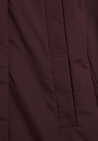 Diep bordeauxkleurige stof met een verticale naad en een horizontale stikseldetails nabij het midden, die lichte kreukels in het materiaal vertoont.