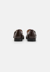 Zapatos de charol marrón con forma redondeada, parte trasera abierta y tacón bajo. Presentan detalles recortados y un lazo decorativo.
