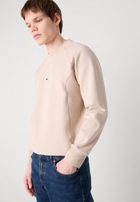 Beige pulóver kerek nyakkivágással, raglán ujjakkal és egy kis címkével a mellrészen. Texturált anyag sötétkék farmernadrággal párosítva.