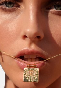 Collier en pendentif en or présentant un design en forme d'œil, orné de cristaux et d'accents verts, suspendu à une fine chaîne en or torsadée.