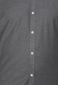 Camisa de botón gris oscuro hecha de tela tejida, con una fila de botones blancos y costuras en contraste a lo largo de la tapeta.