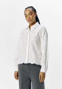 Chemise blanche à boutons avec un design à œillets, ourlet en scallop, manches longues et motifs floraux subtils. Associée à un pantalon gris.
