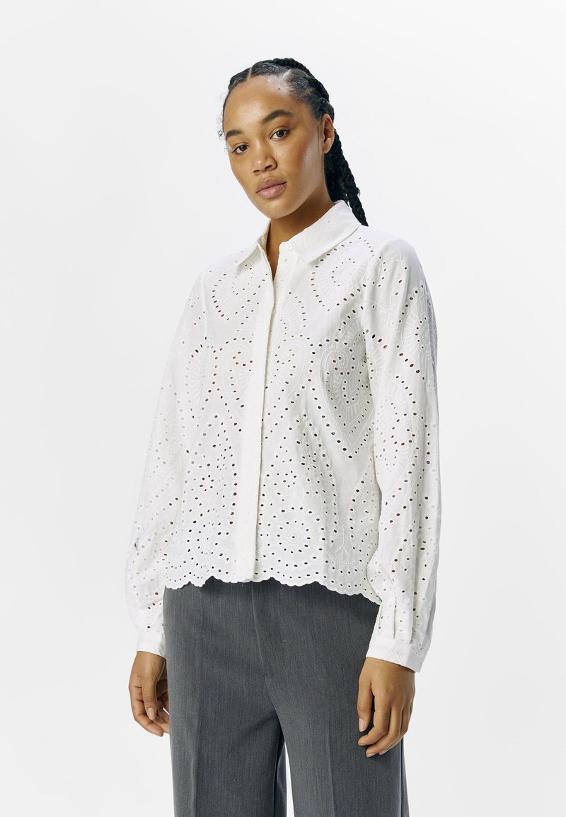 Chemise blanche à boutons avec un design à œillets, ourlet en scallop, manches longues et motifs floraux subtils. Associée à un pantalon gris.