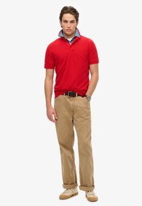 Polo rosso con maniche corte, colletto piegato; chinos beige; sneakers chiare; orologio argentato al polso; design semplice e pulito.