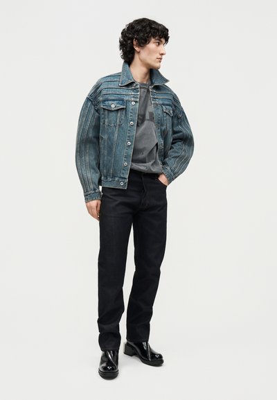 Νέος άνδρας με σγουρά μαλλιά, φοράει μπλε τζιν μπουφάν, γκρι μπλουζάκι με σχέδιο, μαύρο παντελόνι και μαύρα γυαλιστερά μποτάκια, στέκεται μπροστά σε μια μονόχρωμη φόντο.