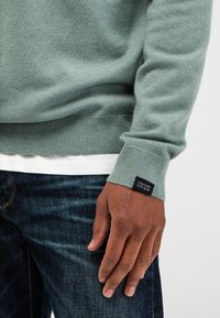 Lichtgroen gebreid sweater met geribde manchetten, voorzien van een zwart etiket "Butcher of Blue" op de mouw, gedragen over een wit T-shirt en donkere jeans.