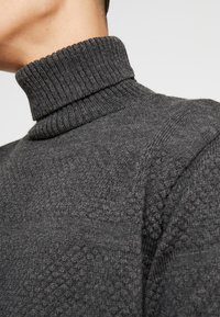 Harmaa, teksturoitu poolopaita, jossa on ribbitekniikalla valmistettu kaulus ja kuviollinen neulos, esittäen lähikuvan kankaan yksityiskohdista ja suunnittelusta.