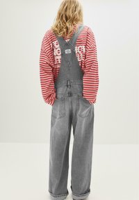 Blond person som bär grå denimoverall och en randig långärmad tröja i röd och vitt, vänd bort mot en neutral vit bakgrund.