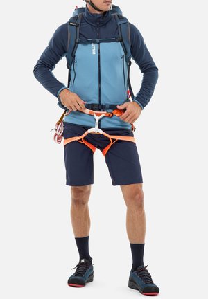 Mann mit Helm, blauem Jacke, Shorts, Wanderschuhen und Rucksack, der einen orangefarbenen Klettergurt um Taille und Oberschenkel anlegt.