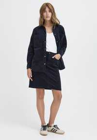 Mornarsko modra kordna srajca-jakna preko belega topa, ujemajoča se mornarsko modra kordna krila in bež superge z črnimi črtami.