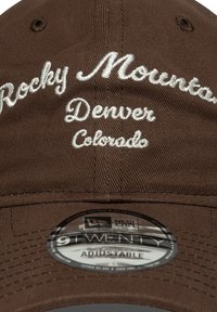 Brūna regulējama cepure ar izšūtu tekstu: "Rocky Mountain," "Denver," un "Colorado" baltā krāsā. Materiāls ir teksturēta auduma.