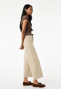 Bruine geribbelde top met korte mouwen, beige wijde pantalon met rafelige zoom en bruine instap schoenen met veters, bekeken vanuit de zijkant.