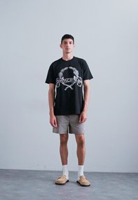 Μαύρο t-shirt με ασημί γραφικό σχέδιο, γκρι καρό σορτς, λευκές κάλτσες και μπεζ παντόφλες, στέκεται σε γκρι δάπεδο.