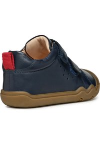 Zapato de cuero azul marino con suela de goma marrón, que presenta una lengüeta de tela roja y perforaciones laterales para transpirabilidad. Cierre con tira de Velcro.