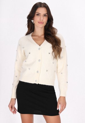 Witte gebreide cardigan met een V-hals, voorzien van ruitpatronen en versierd met heldere, ronde strassaccenten. Draagt over een zwarte rok.