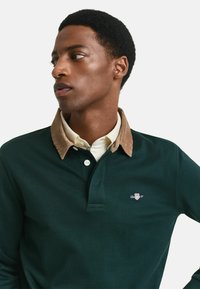 Polo verde scuro con colletto in corduroy beige. Presenta un'apertura con bottoni e un logo ricamato sul lato sinistro del petto. Texture liscia.